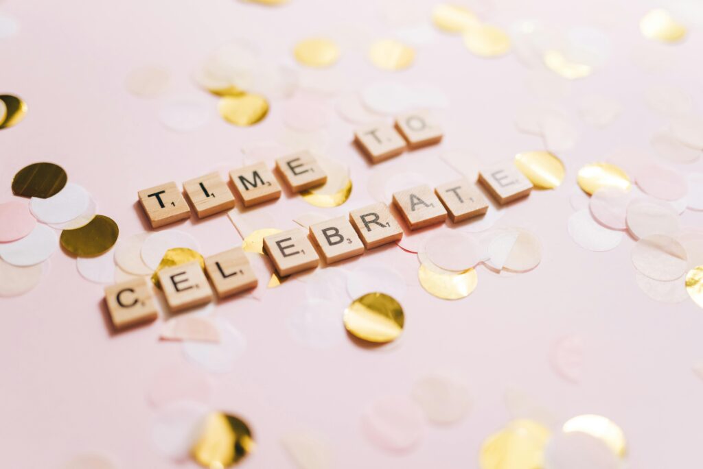 Wooden Scrabble tiles spelling 'Time to Celebrate' on a pink surface with scattered golden confetti.