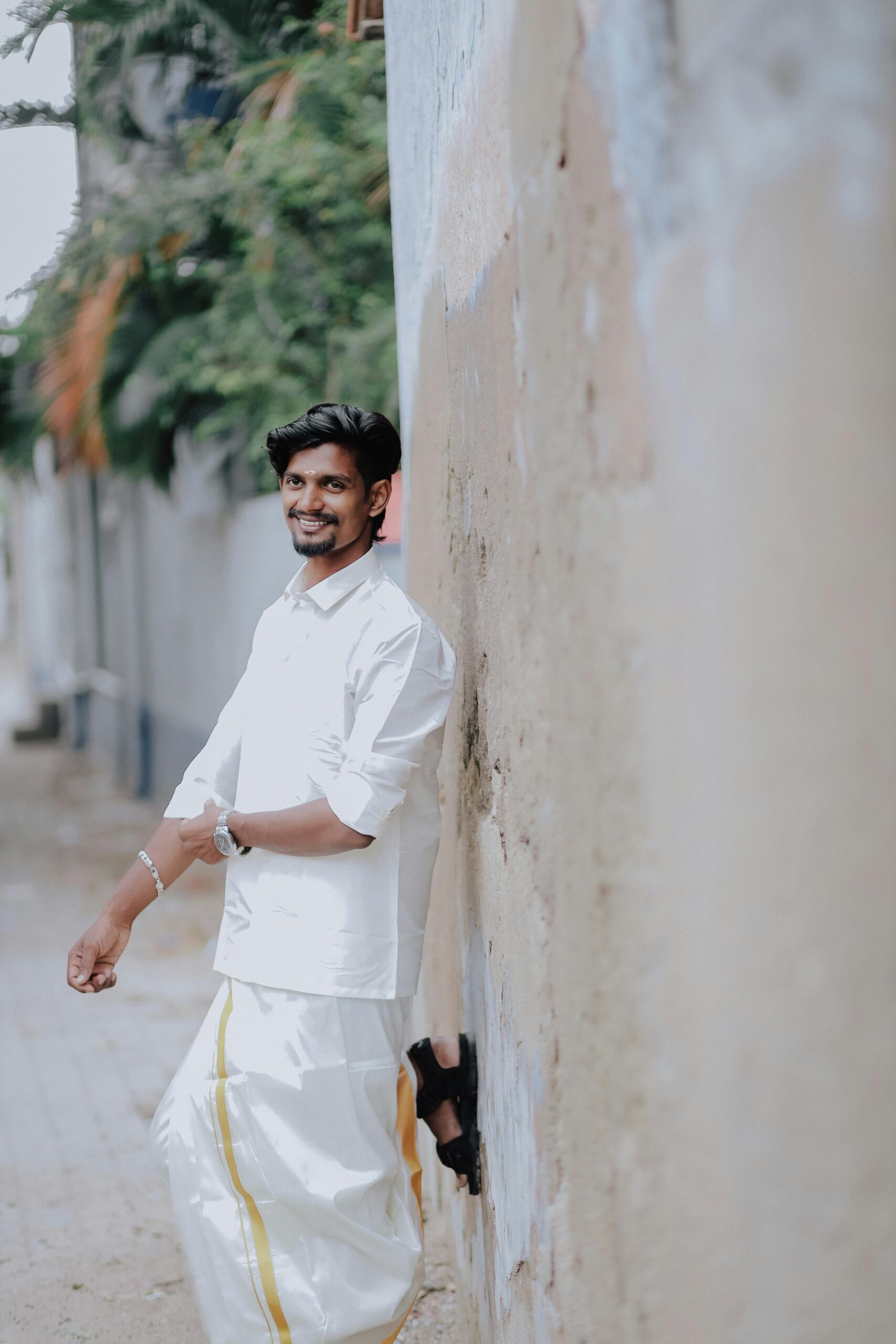 A smiling Indian man wearing traditional attire leaning against a wall outdoors.