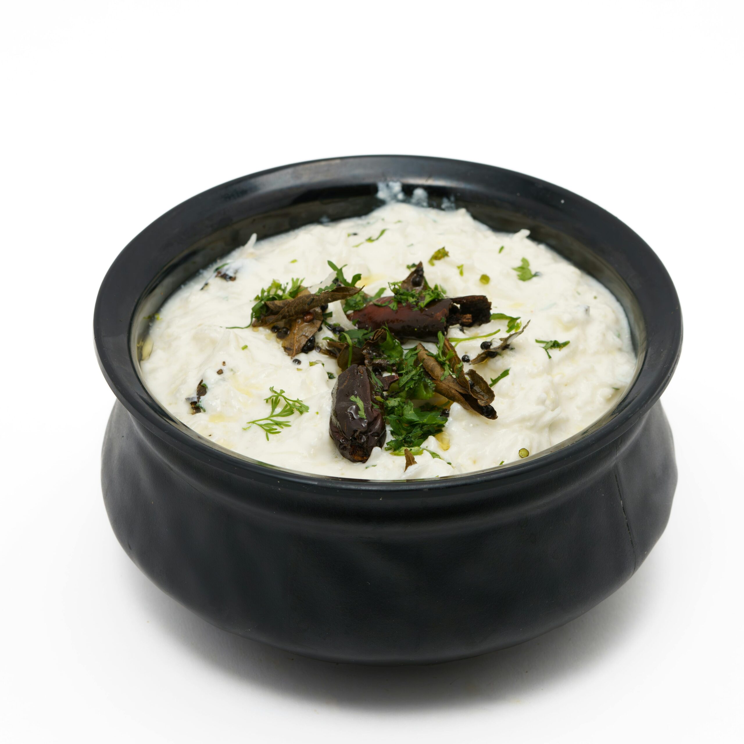 Close-up of creamy curd rice garnished with herbs and dried chilies in a black ceramic bowl.
